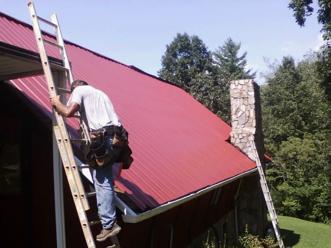 Roofer on a ladder beside a red metal roof and stone chimney — Soffit & Fascia Repair by Roofing Brothers in Cliffside